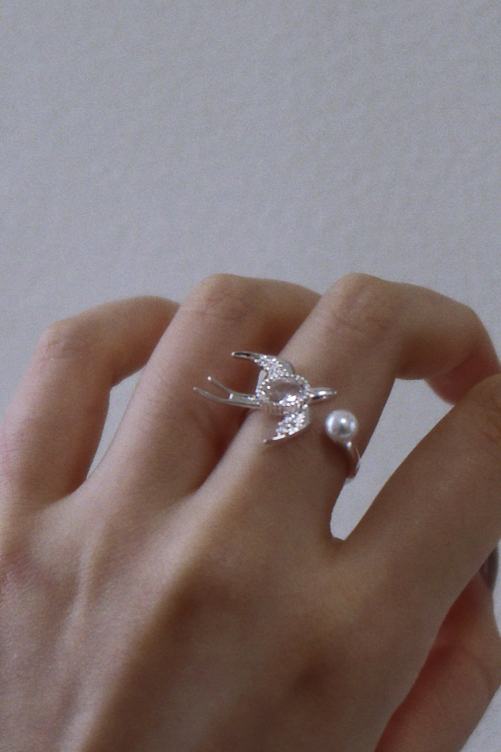 Hand wearing a silver ring with a bird design and a pearl on a plain background