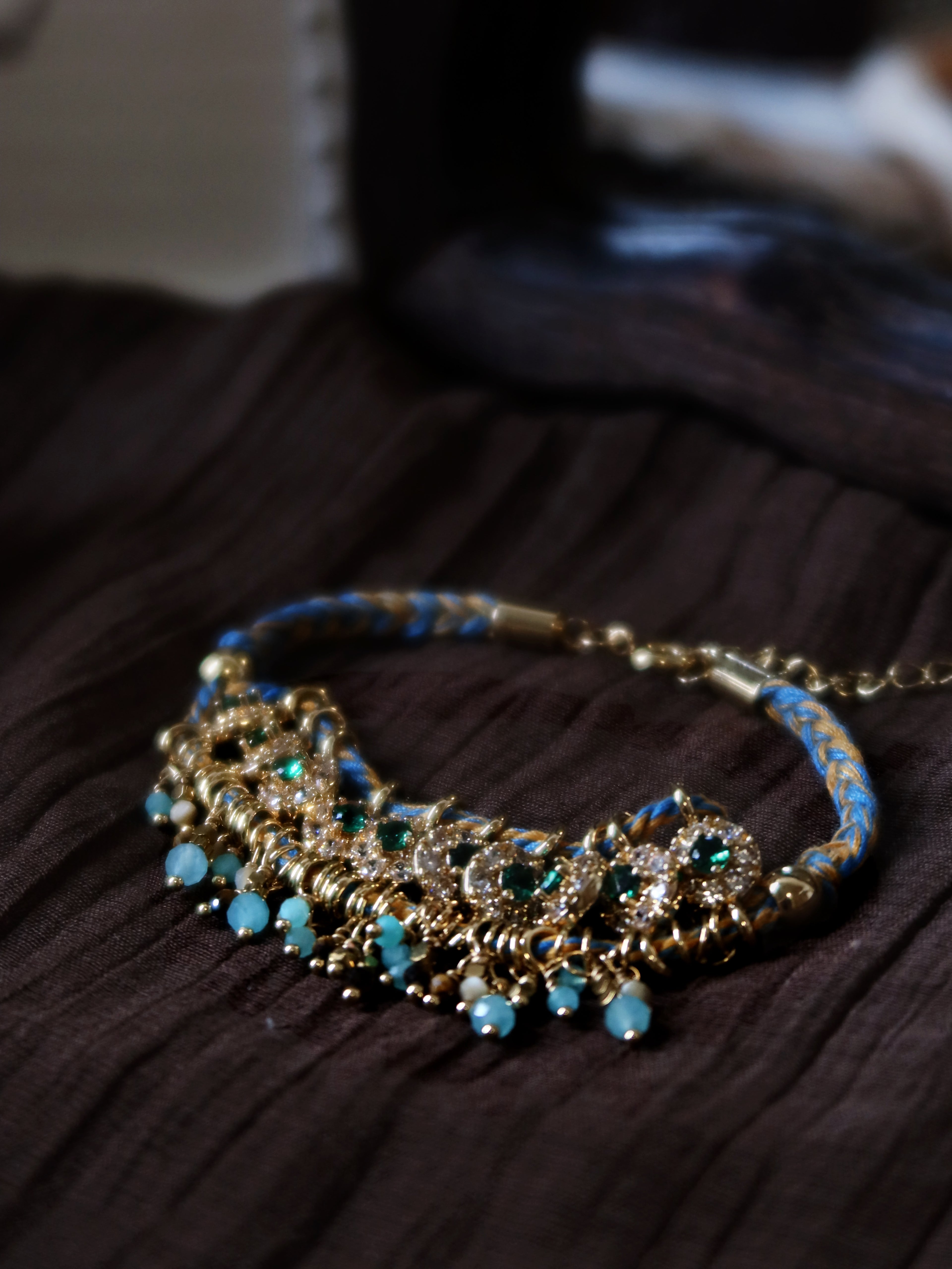 Close-up macro shot of The Peacock’s Muse antique green hand-woven bracelet, showcasing the shimmering green zircon accents, natural tiger's eye beads, and delicate light blue glass tassels under soft natural light.