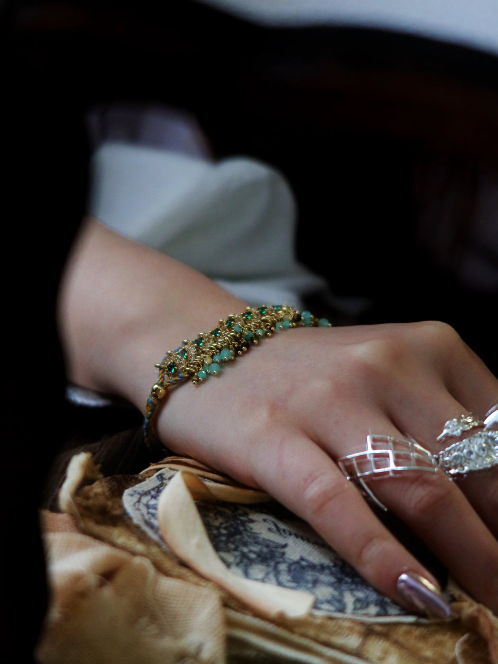 Cinematic vintage aesthetic view of a model wearing The Peacock’s Muse antique green hand-woven bracelet, resting hand on a classical printed prop book.